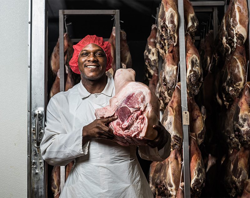 Career Production Volpi employee holding a cured ham with more hams hanging in the background